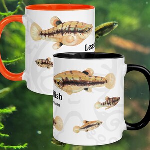 May include: Two white ceramic mugs with fish illustrations. One mug has an orange interior and handle, the other has a black interior and handle. The mugs feature a detailed drawing of a fish with the word "Leas" and "fish" printed on them.