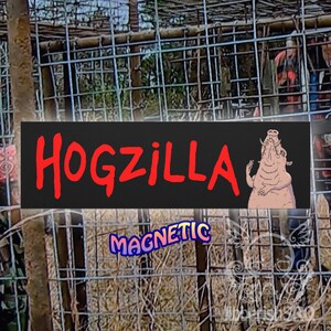 Könnte beinhalten: Ein schwarzes Magnetschild mit dem Wort "HOGZILLA" in roter, stilisierter Schrift. Rechts ist ein Cartoon-Schwein abgebildet. Darunter steht das Wort "MAGNETIC" in blauer, lila und gelber Schrift. Das Schild befindet sich vor einem Drahtzaun.