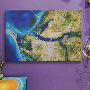 Puede incluir: Una impresión en lienzo rectangular que representa una vista aérea detallada de una zona costera. La obra de arte presenta un océano azul vibrante, masas de tierra verdes y un patrón de cuadrícula que representa el desarrollo urbano. La paleta de colores incluye azules, verdes y amarillos.