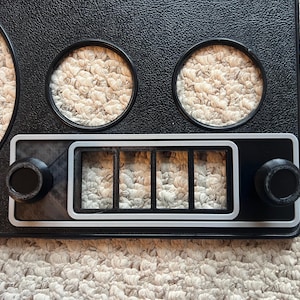 May include: Black dashboard panel with two circular gauge openings, a rectangular section with four rectangular openings, and two round knobs. The panel has a textured surface.