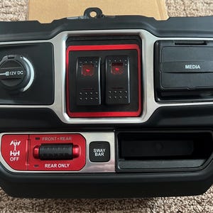 May include: Black automotive control panel with a 12V DC outlet, two illuminated switches, and a media port. Features red accents around the switches and a red panel with text for front/rear and sway bar controls. A small storage area is also included.