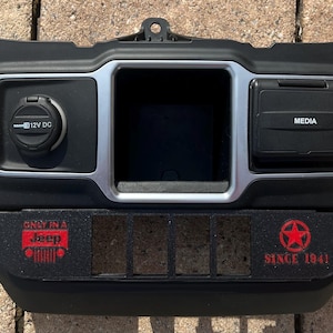 May include: Black car dashboard panel with a 12V DC outlet, media port, and a central compartment. Features the text "ONLY IN A Jeep" and "SINCE 1941" with a star emblem, indicating a Jeep-themed accessory.