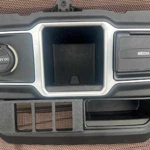 May include: Black automotive console with a 12V DC power outlet, a storage compartment, and a media port labeled "MEDIA". The console also features a section for switches and a small storage area.