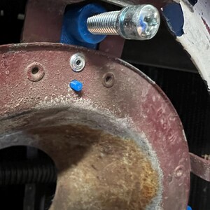 May include: Close-up of a mechanical component featuring a silver bolt secured with a blue plastic spacer. The surrounding metal parts are painted in a faded red color, showing signs of wear and tear.
