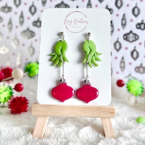 May include: Green and red Christmas ornament earrings. The ornaments are shaped like a traditional Christmas bulb with a green hand reaching out from the top. The earrings are on a white card with the text "Kray Kreations Jewelry".