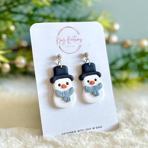May include: A pair of snowman earrings. Each earring features a white snowman with a black top hat, orange nose, and blue scarf. The earrings are on a white card with the text "Klay Kreations Jewelry" and "HANDMADE WITH LOVE IN OHIO".