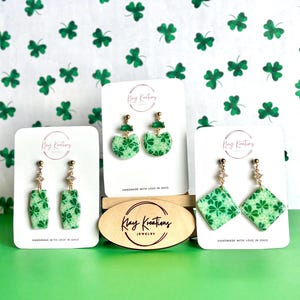 May include: Three pairs of green and white clover-patterned earrings on display. The earrings are on white cards with the text "Klay Kreations Jewelry" and "HANDMADE WITH LOVE IN OHIO". The background is a white surface with green shamrock patterns.