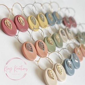 May include: A collection of hoop earrings featuring oval-shaped matte clay pendants in various muted colors, each accented with a small metallic leaf charm. The image includes the text Klay Kreations Jewelry in a circular pink logo design.