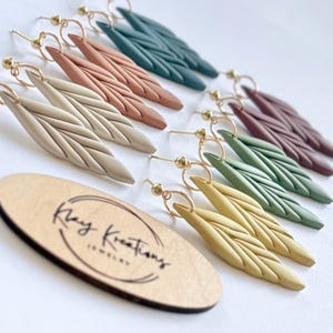 May include: A collection of six pairs of dangle earrings in various colors including beige, terracotta, dark teal, plum, sage green, and mustard yellow. Each earring features a textured, leaf-like design attached to a gold-tone metal hoop.