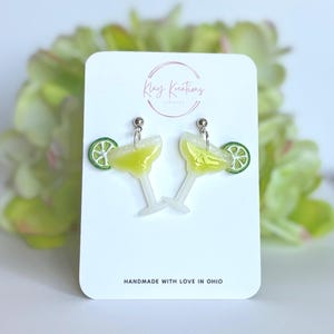 May include: A pair of dangle earrings shaped like margarita glasses with a yellow liquid center, a white rim, and a green lime slice garnish. The earrings are displayed on a white card with the text Kay Kreations Jewelry and Handmade with love in Ohio.