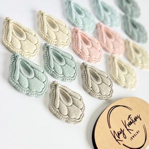 May include: A collection of pastel-colored, wing-shaped jewelry components. The components are in shades of cream, pink, blue, and gray. The components have a textured, detailed design. A wooden sign with the text "Kay Kreations JEWELRY" is in the lower right corner.