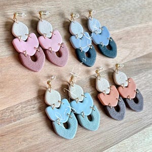 May include: Six pairs of handmade polymer clay earrings in various pastel colors. Each earring features three connected shapes: a top oval, a middle petal shape, and a bottom teardrop with a cutout. Gold-colored earring posts.