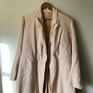 Puede incluir: Un abrigo largo, de doble botonadura, de color beige con un patrón a cuadros. El abrigo tiene una percha de madera marrón y está colgado en una pared de color verde claro.