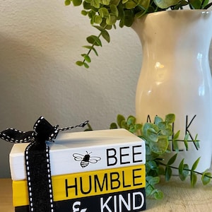 Puede incluir: Una pila de tres bloques de madera decorativos con las palabras "BEE HUMBLE & KIND" en blanco y negro. Una cinta negra con costuras blancas está atada alrededor de los bloques. Un gráfico de abeja está en el bloque superior. Un jarrón blanco con vegetación está en el fondo.