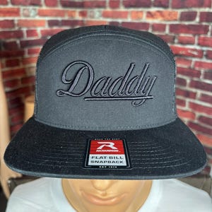 May include: A black snapback hat with a flat bill and a mesh back. The front panel features the word Daddy embroidered in black script font. A rectangular sticker on the bill displays the Richardson logo and the text Flat Bill Snapback.
