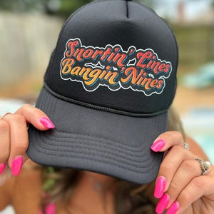 May include: Black trucker hat with a colorful embroidered logo that says "Snortin' Lines Bangin' Nines".