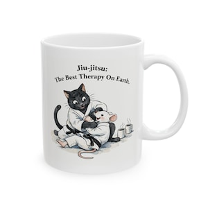 Puede incluir: Taza de cerámica blanca con una ilustración de dibujos animados de un gato negro y un ratón blanco con uniformes de Jiu-jitsu. La taza tiene el texto "Jiu-jitsu: The Best Therapy On Earth." También se representan dos tazas de café pequeñas.