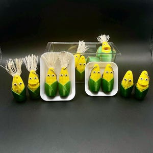 Op de afbeelding: Een verzameling maïsvormige sleutelhangers met lachende gezichten. De sleutelhangers zijn geel met groene schillen en witte kwastjes. Sommige worden weergegeven in witte bakjes, andere in een doorzichtige container, allemaal op een donkere ondergrond.