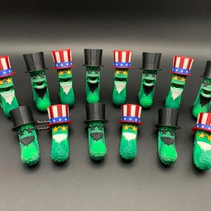 May include: A group of green, cylindrical figurines with white beards and smiling faces. Some wear black top hats, while others have red, white, and blue Uncle Sam hats. The figurines are displayed on a dark surface.