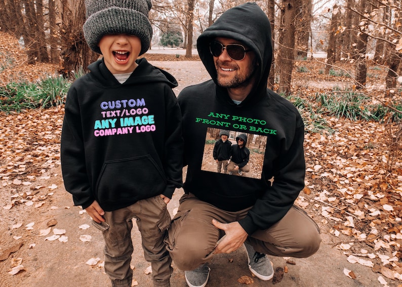 May include: Two people wearing black hooded sweatshirts with custom text and images. The text on the sweatshirt on the left says "CUSTOM TEXT/LOGO ANY IMAGE COMPANY LOGO". The text on the sweatshirt on the right says "ANY PHOTO FRONT OR BACK".