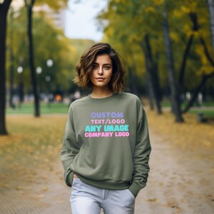 May include: An olive green crewneck sweatshirt featuring the text CUSTOM TEXT/LOGO ANY IMAGE COMPANY LOGO printed in light purple, pink, and cyan colors on the front chest area. The garment has long sleeves and a ribbed neckline and cuffs.