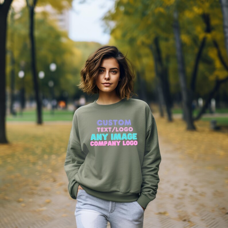 May include: An olive green crewneck sweatshirt featuring the text CUSTOM TEXT/LOGO ANY IMAGE COMPANY LOGO printed in light purple, pink, and cyan colors on the front chest area. The garment has long sleeves and a ribbed neckline and cuffs.