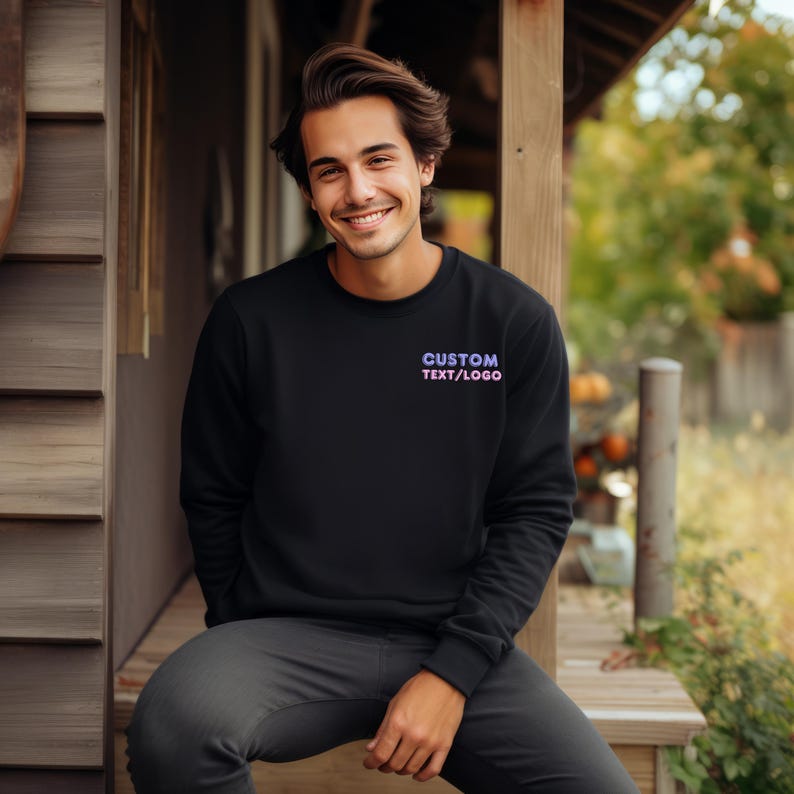 May include: A black long-sleeve crewneck sweatshirt featuring the text CUSTOM TEXT/LOGO in a purple and pink gradient font on the upper left chest area. The sweatshirt has a ribbed neckline, cuffs, and hem, providing a clean and simple look.