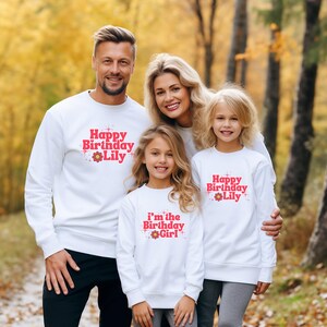 May include: White crewneck sweatshirts featuring red text and a flower graphic. Two sweatshirts display the text Happy Birthday Lily with star accents, while one sweatshirt displays the text i'm the Birthday Girl with star accents.