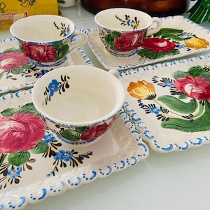 Puede incluir: Un juego de tazas de té de cerámica y bandejas rectangulares a juego. Las tazas y las bandejas son blancas con un diseño floral con rosas rojas, hojas verdes y detalles azules. Las bandejas tienen un borde festoneado con puntos azules.
