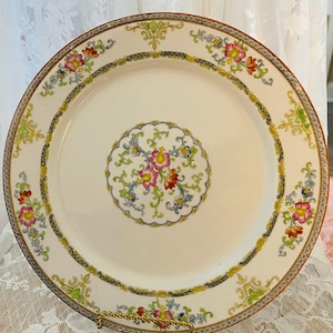 May include: A round, white ceramic plate with a floral design. The plate features a central floral medallion and a border with repeating floral and scroll motifs in pink, yellow, blue, and green. The rim has a gold-coloured trim.