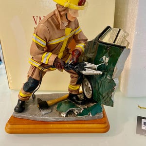 Puede incluir: Figura de un bombero con uniforme beige y casco amarillo, trabajando en un coche verde dañado. El bombero sostiene una herramienta, con un hacha cerca. La figura está montada sobre una base de madera.
