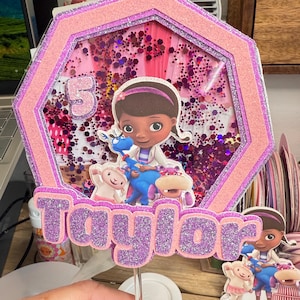 May include: A custom birthday cake topper featuring the character Doc McStuffins, the number 5, and the name "Taylor" in glittery purple letters. The topper has a pink and purple hexagonal frame with a clear center filled with confetti.