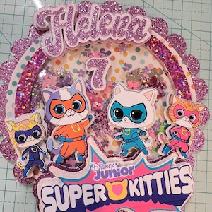 Puede incluir: Un adorno para pastel de purpurina rosa y morado con el nombre "Helena" y el número "7" en él. El adorno también presenta el logotipo de Disney Junior y las palabras "Super Kitties". Hay cuatro gatos de dibujos animados vestidos de superhéroes en el adorno.