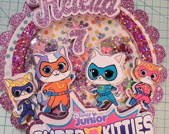Op de afbeelding: Een roze en paarse glittertaarttopper met de naam "Helena" en het nummer "7" erop. De topper heeft ook het Disney Junior logo en de woorden "Super Kitties". Er zijn vier cartoonkatten verkleed als superhelden op de topper.