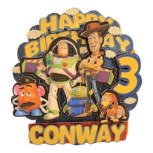 May include: A colourful cake topper featuring characters from the Toy Story film franchise. The topper includes Woody, Buzz Lightyear, Mr. Potato Head, and a green alien. The topper also includes the words "Happy Birthday" and the number "3" in yellow and blue. The name "Conway" is written in yellow letters on a red background.