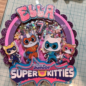 May include: A pink and purple cake topper with a sparkly background. The topper features the name "Ella" and the number "7" in pink glitter. There are four cartoon cats dressed as superheroes with the text "Disney Junior Super Kitties".