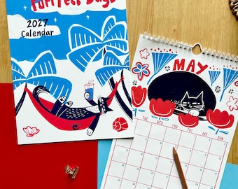 May include: A 2027 wall calendar titled Purrfect Days featuring a black cat in a red hammock on the cover, and an open calendar page for May with a black cat illustration, red flowers, and a wooden pencil resting on the grid.