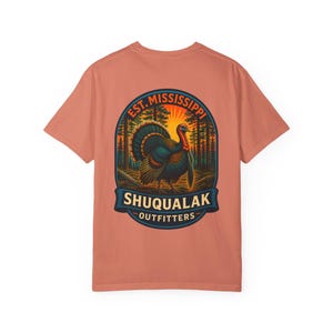 May include: A coral-colored short-sleeve t-shirt featuring a graphic on the back. The design shows a turkey in a forest at sunset, enclosed in an oval frame with the text EST. MISSISSIPPI, SHUQUALAK, and OUTFITTERS in bold lettering.