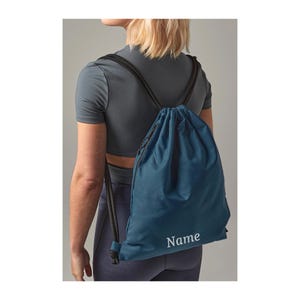 Könnte beinhalten: Eine dunkelblaue Sporttasche mit schwarzen Bändern und einem weißen, gestickten Namen. Die Tasche wird von einer Person getragen.