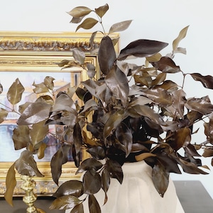 May include: A bouquet of dried brown leaves in a cream-coloured vase. The vase is sitting on a dark surface in front of a gold-framed picture.