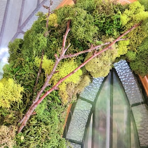 May include: A green moss wreath with brown branches woven throughout. The wreath is hanging on a wooden door with a stained glass window.