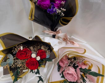May include: Three bouquets of preserved roses wrapped in decorative paper. The bouquets are arranged in a triangular shape. The roses are pink, red, and purple. The bouquets are tied with a ribbon.