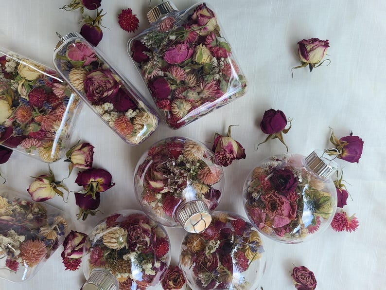 May include: A collection of seven clear glass ornaments filled with dried flowers and greenery. The ornaments are various shapes and sizes, including round, square, and rectangular. The flowers are primarily shades of pink, purple, and white. The ornaments are arranged on a white surface.