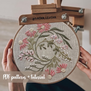 May include: A round embroidery hoop with a floral design. The embroidery features pink and white flowers, green leaves, and white bead accents. The text "PDF pattern + tutorial" is at the bottom. The wooden embroidery stand is visible at the top.