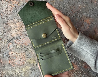 May include: A dark green leather wallet with yellow stitching and a small pocket on the front. The wallet is open and has multiple card slots and a billfold. The wallet has a small tree logo embossed on the inside.