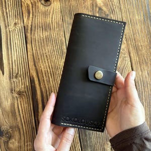 Peut inclure: Un long portefeuille rectangulaire en cuir noir au fini mat, doté d'une fermeture à bouton-pression en laiton vieilli et de coutures contrastantes claires sur les bords. Le nom SOKOLOVSKY est embossé sur la partie inférieure avant.