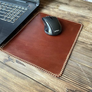 Puede incluir: Un mousepad de cuero marrón con un borde cosido y un ratón de ordenador negro. El mousepad es rectangular y está sobre una superficie de madera junto a un portátil.