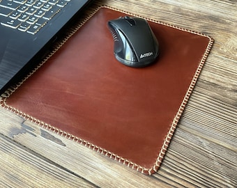 May include: A brown leather mousepad with a stitched border and a black computer mouse. The mousepad is rectangular and sits on a wooden surface next to a laptop.