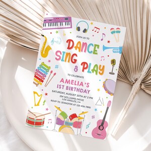 May include: A colorful invitation with the text "Dance, Sing & Play" and musical instrument illustrations. The invitation is for Amelia's 1st birthday, with the date, time, and location details. The invitation is on a white plate with a dried palm leaf.