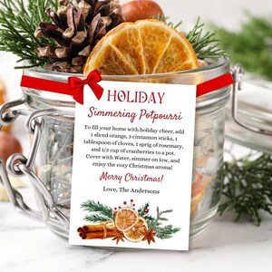 May include: A glass jar filled with dried orange slices, cinnamon sticks, pine cones, and other holiday-themed ingredients. A red ribbon with a bow is tied around the jar. A white card with red text reads "HOLIDAY Simmering Potpourri" and includes instructions for making a holiday-scented simmer pot. The card also includes a watercolor illustration of holiday-themed ingredients.